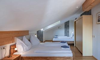 A cozy bedroom with two beds and wooden details. The roof is slanted, and there is plenty of natural light coming through the windows.