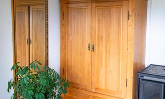 Large wooden wardrobe, full-length mirror, and a green indoor plant create a warm, homely atmosphere. Suitcases and clothes are well stored, and the room remains tidy.