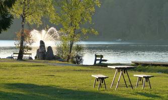 A quiet park by the lake with benches and a water fountain. The sun shines through the trees and reflects on the water's surface.