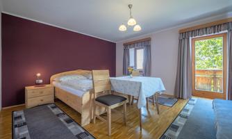 A cozy room with a bed, a dining table, and chairs. The room has large windows that let in plenty of daylight.