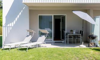 A bright garden area with two loungers and a sun umbrella. In the background, there is a dining table and a grill.