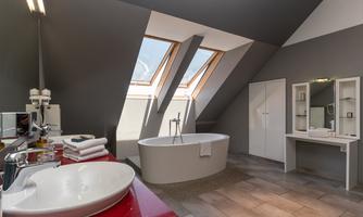 A modern bathroom with slanted windows and a freestanding bathtub. The walls are gray, and there is a tasteful decor with a sink and a large mirror.