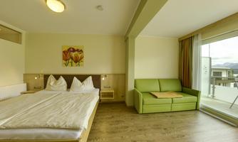 A comfortable hotel room with a double bed and a green sofa. Large windows offer plenty of natural light and lead to a balcony.