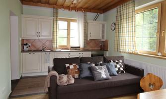 A cozy kitchen with a modern sofa and large windows. The room is bright and inviting with wooden details and checkered curtains.