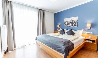 A modern bedroom with a large bed and blue walls. The room is bright and friendly, with curtains that let in plenty of daylight.