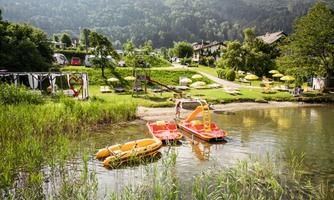 A calm lake with two red pedal boats and green shore vegetation. In the background are meadows and trees that create an idyllic landscape.