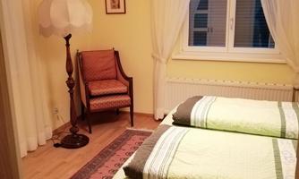 A cozy bedroom with two single beds and a chair. The room is decorated in warm shades of yellow and has large windows with curtains.