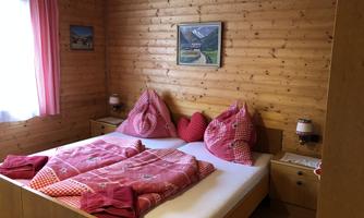 A cozy bedroom with wooden walls and a large bed. The ceiling and pillows are in shades of red and pink.