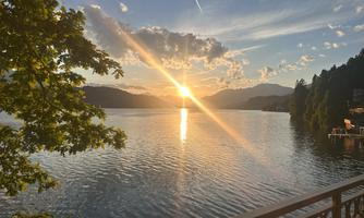 A picturesque sunset over a calm lake. The sun reflects in the water, surrounded by trees and mountains.