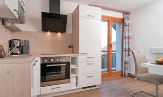 A modern kitchen with bright wood and white cabinets. It features a stove, a coffee machine, and a balcony with a window.