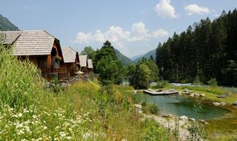 An idyllic landscape with wooden cabins by the water. Surrounded by green meadows and tall trees under a blue sky.