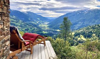 A viewing terrace with comfortable chairs and a view of green valleys and mountains. The sky is cloudy, and the landscape is idyllic and peaceful.