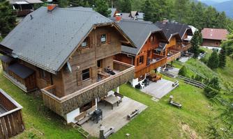 A series of cozy wooden chalets in a green environment. The houses have large balconies and are surrounded by trees.