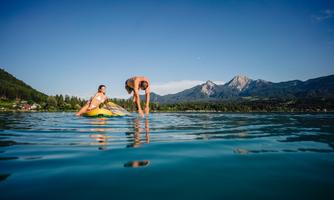 Two people in the water, one on a swim ring and the other bending down. In the background, beautiful mountains and a clear blue sky can be seen.