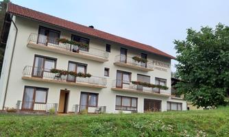 A modern guesthouse with several balconies and flower boxes. It is situated in a green environment, surrounded by trees.