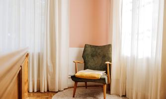 A cozy armchair is situated in a bright room with white curtains and a soft pink wall. A yellow blanket lies on the armchair, creating an inviting atmosphere.
