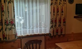 A cozy dining area with wooden chairs and tables. Flowering curtains hang in front of a window.