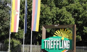 A welcome sign in Treffling with sun symbolism and various flags. The surroundings are green and wooded.