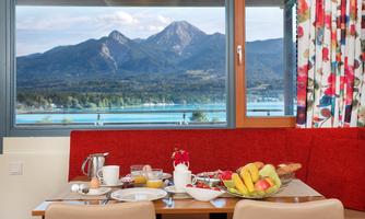 A cozy breakfast table with fresh fruits, pastries, and drinks. In the background, a magnificent mountain landscape and a clear lake can be seen.