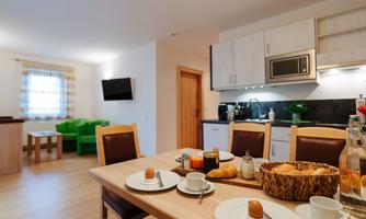 A cozy kitchen with a dining area set for breakfast. In the background, a bright living room with a green sofa and a television can be seen.