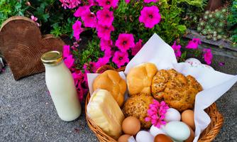 A basket filled with fresh rolls, cookies, eggs, and milk is in front of colorful flowers. The scene conveys a cozy, rural atmosphere.