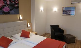 A modern hotel room with a double bed and red decorative pillows. There is a cozy armchair and nice wall decoration.