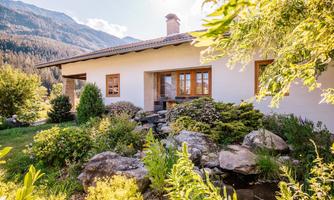 A beautiful house surrounded by a garden with colorful plants and stones. In the background, the mountains and a clear sky can be seen.