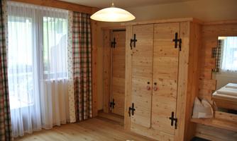 A cozy room with a wooden wardrobe and a large window. Bright curtains and wooden flooring create a warm atmosphere.