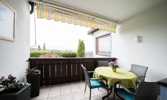 A cozy balcony with a table and chairs. The sunshade provides shade and there are some plants in the background.