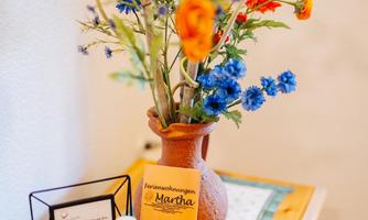 A beautiful bouquet of blue and orange flowers stands in a clay vase. Next to it, a document and glasses with food can be seen on a piece of wood.