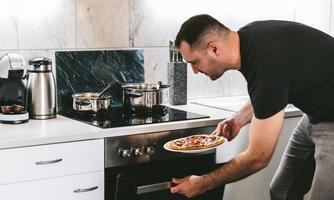 A man in a modern kitchen is taking a pizza out of the oven. Several pots are on the stove.