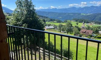 A picturesque landscape with green meadows and mountains in the background. The view from a balcony shows a peaceful, rural setting.
