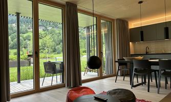 A modern living room with large windows overlooking a green landscape. There is a dining table, chairs, and cozy seating cushions on a carpet.