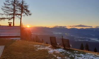 A tranquil mountain landscape with a sunset in the background. Two loungers are placed on the meadow, surrounded by pine trees.