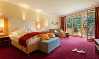 An elegant hotel room with a large bed and a cozy seating area. The balcony offers a view of the green countryside.