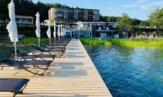 A wooden terrace leads to a tranquil body of water. In the background, there is a hotel with landscaped shore areas.