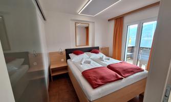 A modern bedroom with a double bed and red pillows. Large windows provide plenty of daylight and offer a view of a balcony.