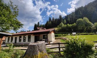 A cozy house surrounded by nature, with green meadows and trees. The sky is partly cloudy and the landscape is hilly.