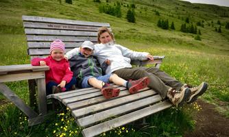 A woman is sitting with two children on a large wooden bench in a green landscape. In the background, gentle hills and trees can be seen.