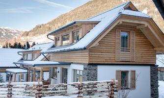 A modern wooden house in the snow, surrounded by mountains. The facade combines wood and stone, with a veranda and shingle roof.