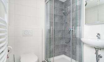 A modern bathroom with a glass shower cabin. There is a toilet and a small sink on the wall.
