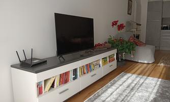 A living room with a television on a white TV shelf. The shelf contains books and there is a cozy armchair as well as plants in the room.