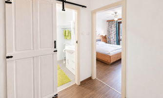 A bright room with an open door to a bathroom and another door to a bedroom. The walls are light, the floor is wooden, and there are some decorative elements.