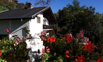 A colorful flower landscape with red geraniums and a small garden figure. In the background, a white house is visible against a bright blue sky.