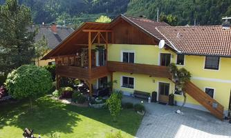 A yellow house with a large balcony and a green lawn surrounded by trees. In the background, gentle hills can be seen.
