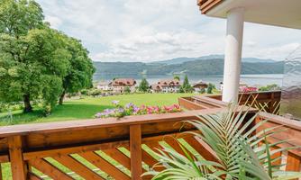 A beautiful view from a balcony overlooking a green meadow and the lake. In the background, mountains and some houses can be seen.