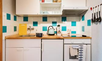 A modern kitchen with blue and white tiles. In the foreground, there is a kettle, a sink, and various kitchen appliances.