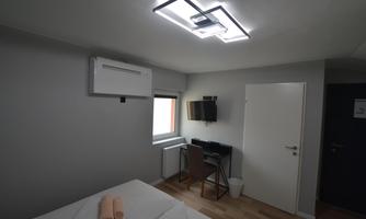 A modern bedroom with wooden flooring and minimalist furnishings. There is a bed, a desk, and an air conditioning unit.