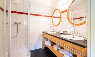 A modern bathroom with two sinks and large mirrors. It features a shower and a stylish wooden shelf.