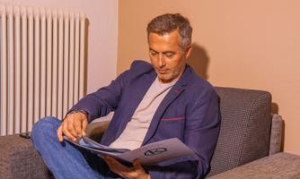A man is sitting on a gray sofa and reading a brochure. He is wearing a casual outfit and is focused on the pages.